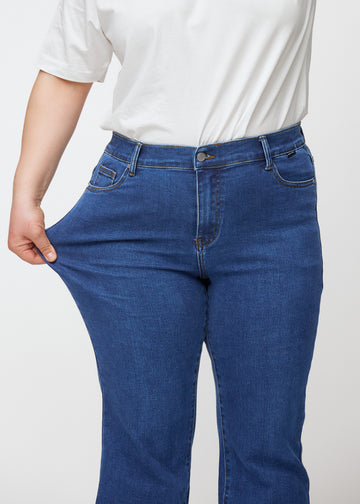 Dunkelblaue Mid-Rise-Bootcut-Jeans in der Farbe Oceans. Ein Plus-Size-Model zieht am Jeansstoff am Oberschenkel, um den Stretch zu zeigen.