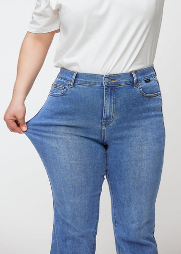 Blaue Mid-Rise-Bootcut-Jeans in der Farbe Rivers. Ein Plus-Size-Model zieht am Jeansstoff am Oberschenkel, um den Stretch zu zeigen.