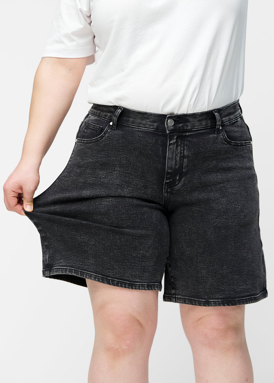 Plus-size model strækker de løse, mørkegrå denim shorts i farven Shadows ved hoften for at vise materialets stretch.