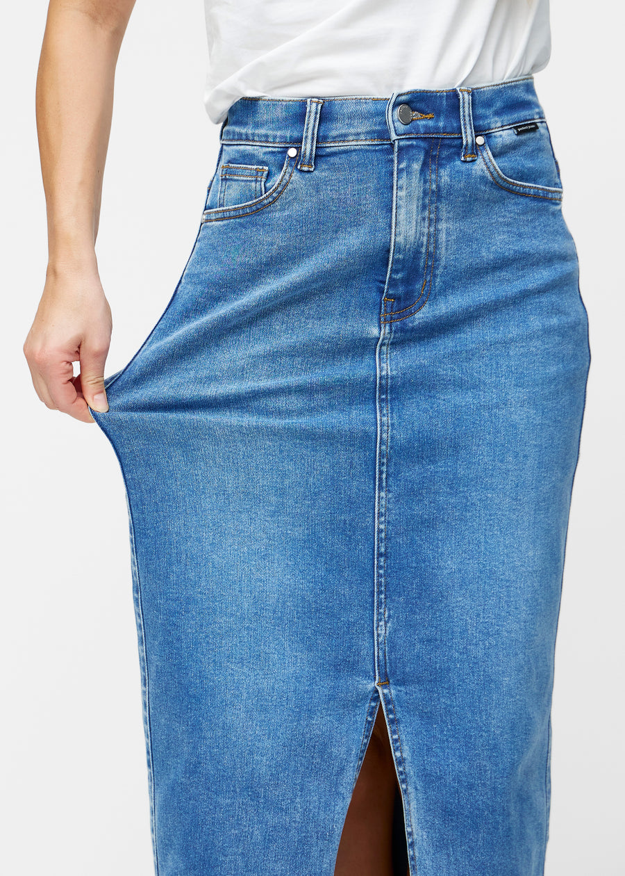 Model strækker den lange, klassisk blå denimnederdel i farven Rivers ved hoften for at vise materialets stretch.