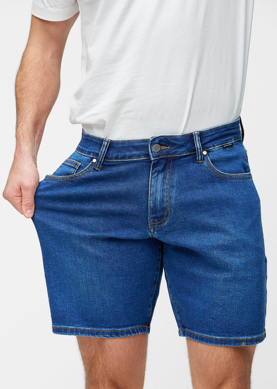Modell dehnt dunkelblaue Denim-Shorts für Herren in der Farbe Oceans am Oberschenkel, um den Stretch des Materials zu zeigen.