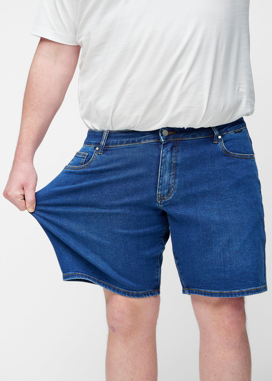 Plus-Size-Modell dehnt dunkelblaue Denim-Shorts für Herren in der Farbe Oceans am Oberschenkel, um den Stretch des Materials zu zeigen.