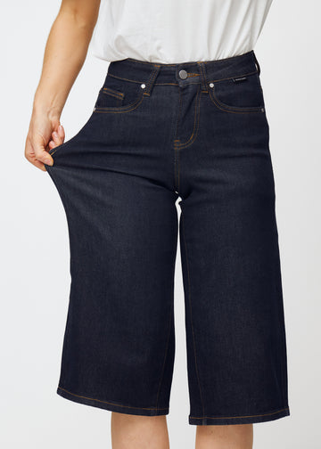 Model dehnt tief dunkelblaue Denim-Capris für Damen in der Farbe Midnights am Oberschenkel, um den Stretch des Materials zu zeigen.