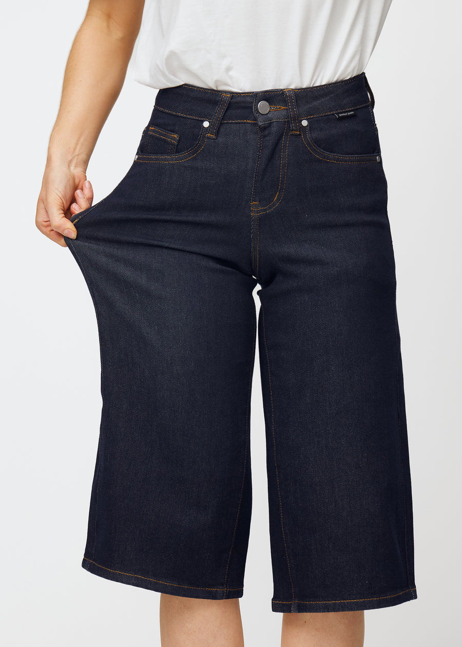 Model dehnt tief dunkelblaue Denim-Capris für Damen in der Farbe Midnights am Oberschenkel, um den Stretch des Materials zu zeigen.