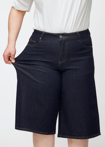 Plus-Size-Model dehnt tief dunkelblaue Denim-Capris für Damen in der Farbe Midnights am Oberschenkel, um den Stretch des Materials zu zeigen.