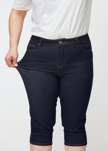 Plus-Size-Model dehnt tief dunkelblaue Denim-Capris für Damen in der Farbe Midnights am Oberschenkel, um den Stretch zu zeigen.