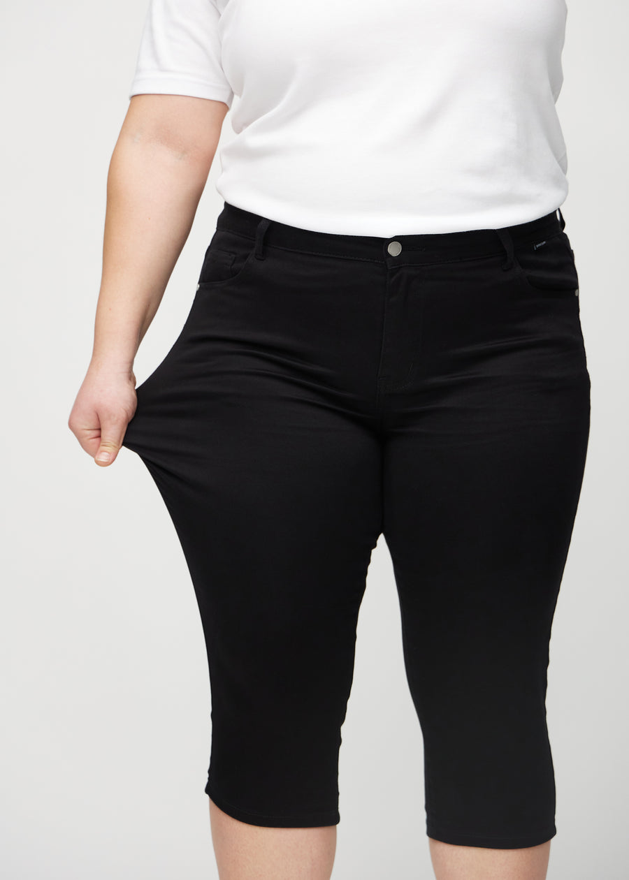 Skinny-Capris für Damen in schwarzem Jeansstoff, Farbe Ravens. Ein Plus-Size-Model dehnt den Stoff, um den Stretch zu zeigen.
