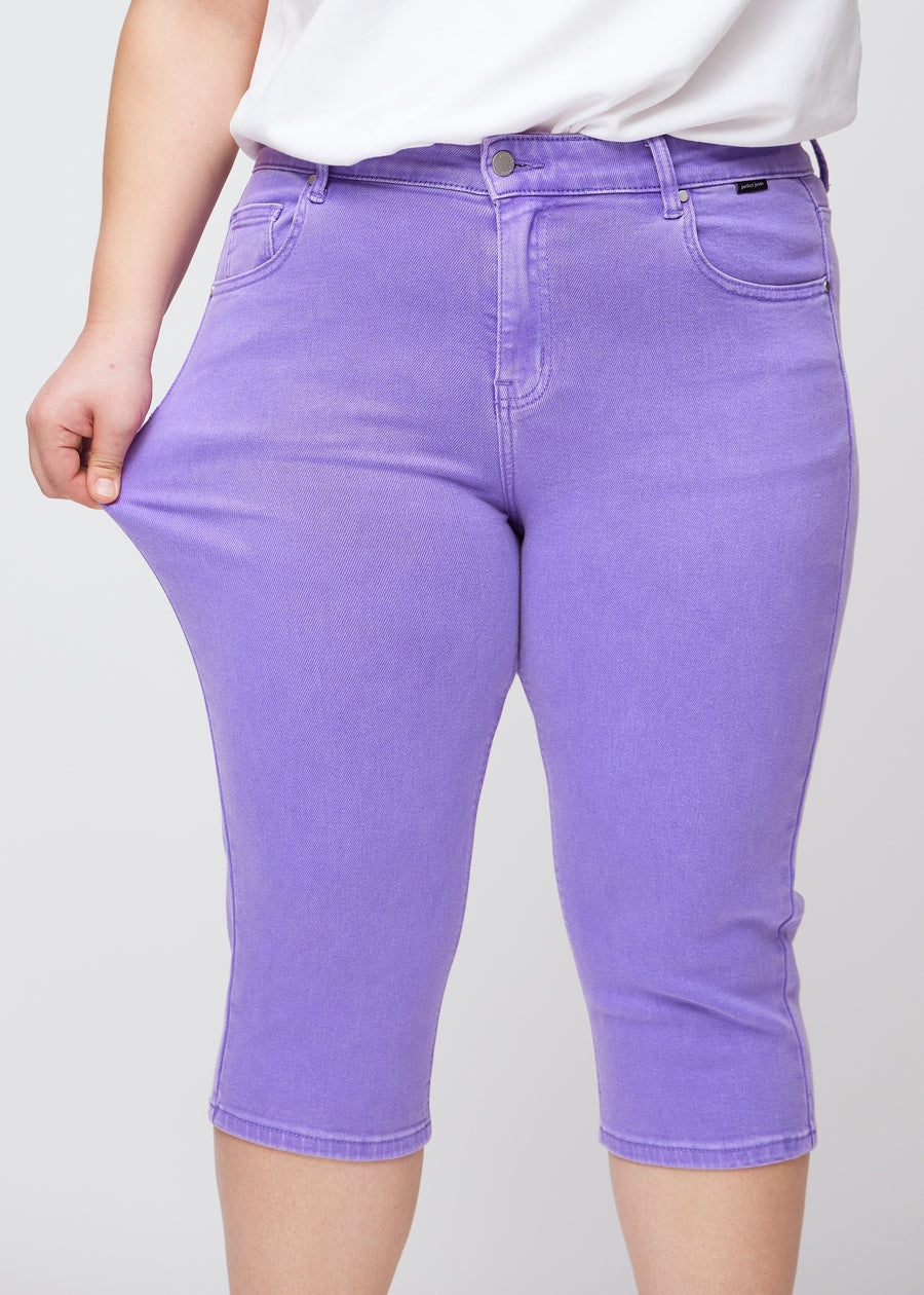 Slim-Capris für Damen aus pastellviolettem Jeansstoff, Farbe Lavenders. Ein Plus-Size-Model dehnt den Stoff am Oberschenkel, um die Flexibilität zu zeigen.