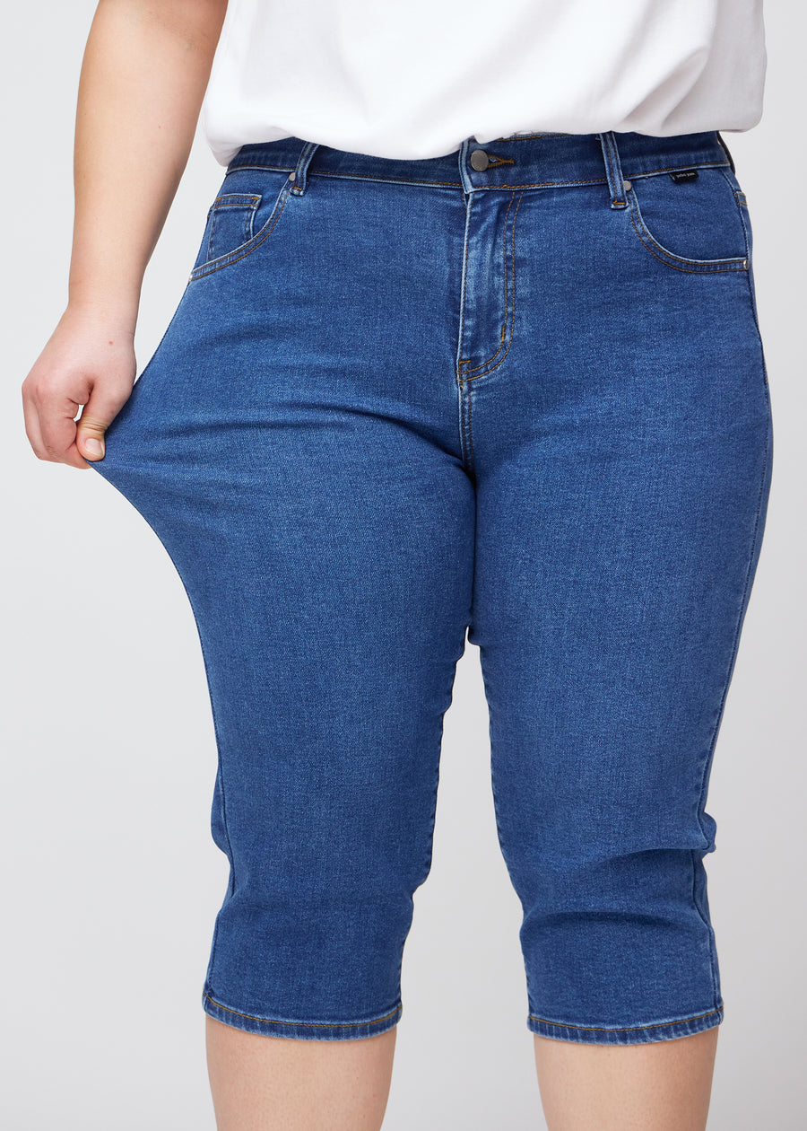 Ein Plus-Size-Model zeigt den Stretch, indem es die Capris am Oberschenkel zieht, Fokus auf Komfort und Flexibilität im dunkelblauen Denim.