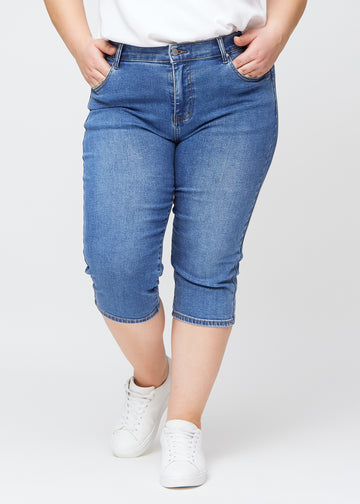 Slim-Capris für Damen aus blauem Denim, Farbe Rivers. Vorderansicht an einem Plus-Size-Model, figurbetonter Schnitt bis knapp unter das Knie.