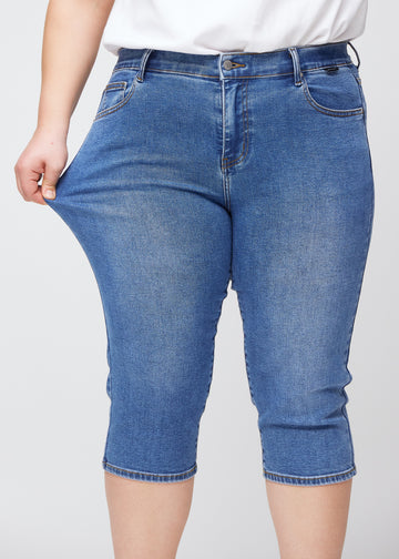 Slim-Capris für Damen aus blauem Denim, Farbe Rivers. Ein Plus-Size-Model dehnt den Stoff am Oberschenkel, um die Flexibilität zu zeigen.