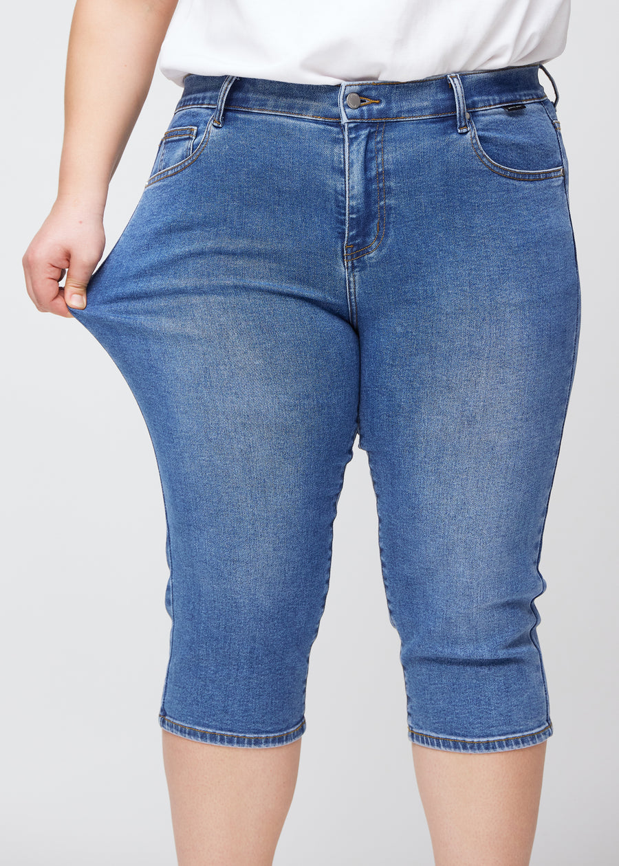 Slim-Capris für Damen aus blauem Denim, Farbe Rivers. Ein Plus-Size-Model dehnt den Stoff am Oberschenkel, um die Flexibilität zu zeigen.