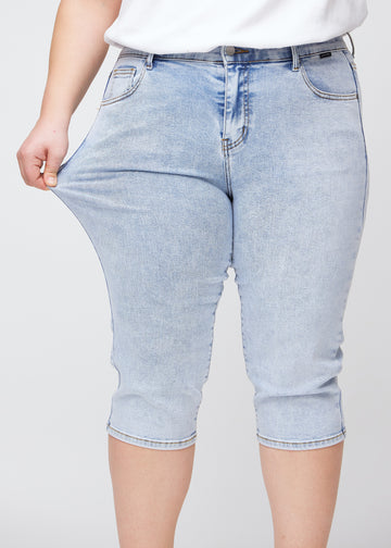 Slim-Capris für Damen aus hellblauem Denim, Farbe Waves. Ein Plus-Size-Model dehnt den Stoff am Oberschenkel, um die Flexibilität zu zeigen.