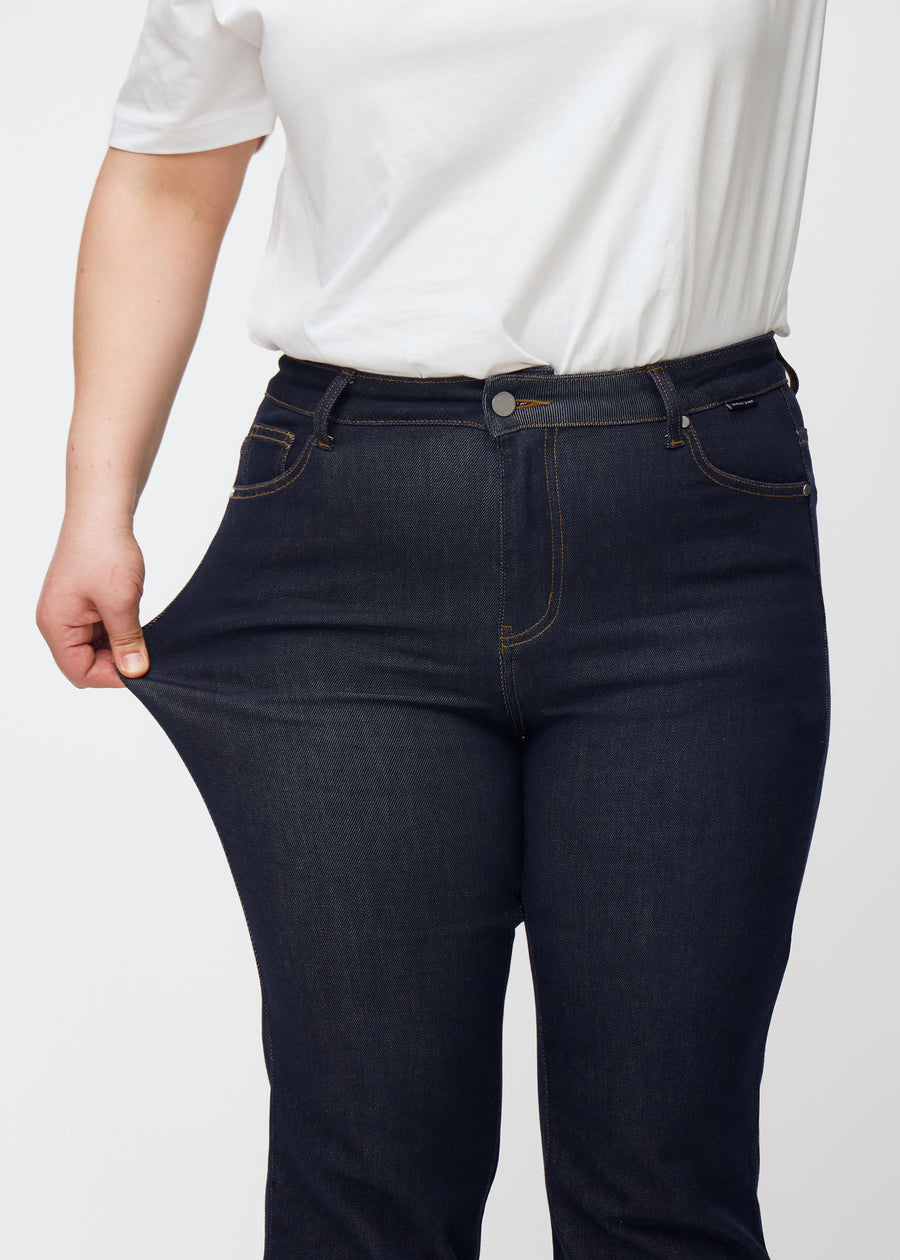 Ein Plus-Size-Model dehnt die tief dunkelblaue Denim jeans für Damen in der Farbe Midnights am Oberschenkel, um die Dehnbarkeit des Materials zu zeigen.