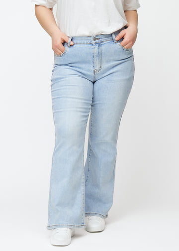 Hellblaue Mid-Rise-Bootcut-Jeans in der Farbe Waves. Vorderansicht an einem Plus-Size-Model, an Hüfte und Oberschenkeln anliegend mit leicht ausgestelltem Bein.