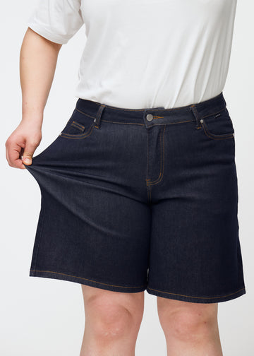 Plus-Size-Model dehnt tief dunkelblaue Denim-Shorts für Damen in der Farbe Midnights am Oberschenkel, um den Stretch zu zeigen.