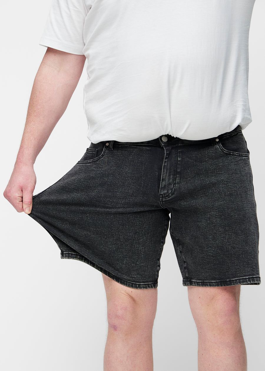 Plus-Size-Modell dehnt dunkelgraue Denim-Shorts für Herren in der Farbe Shadows am Oberschenkel, um den Stretch des Materials zu zeigen.