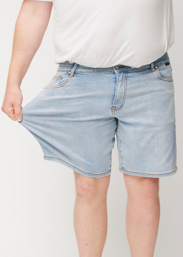 Das Plus-Size-Model dehnt hellblaue Denim-Shorts für Herren in der Farbe Waves am Oberschenkel, um den Stretch des Materials zu zeigen.