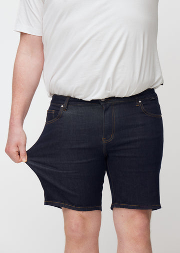 Plus-Size-Model dehnt tief dunkelblaue Denim-Shorts für Herren in der Farbe Midnights am Oberschenkel, um den Stretch zu zeigen.