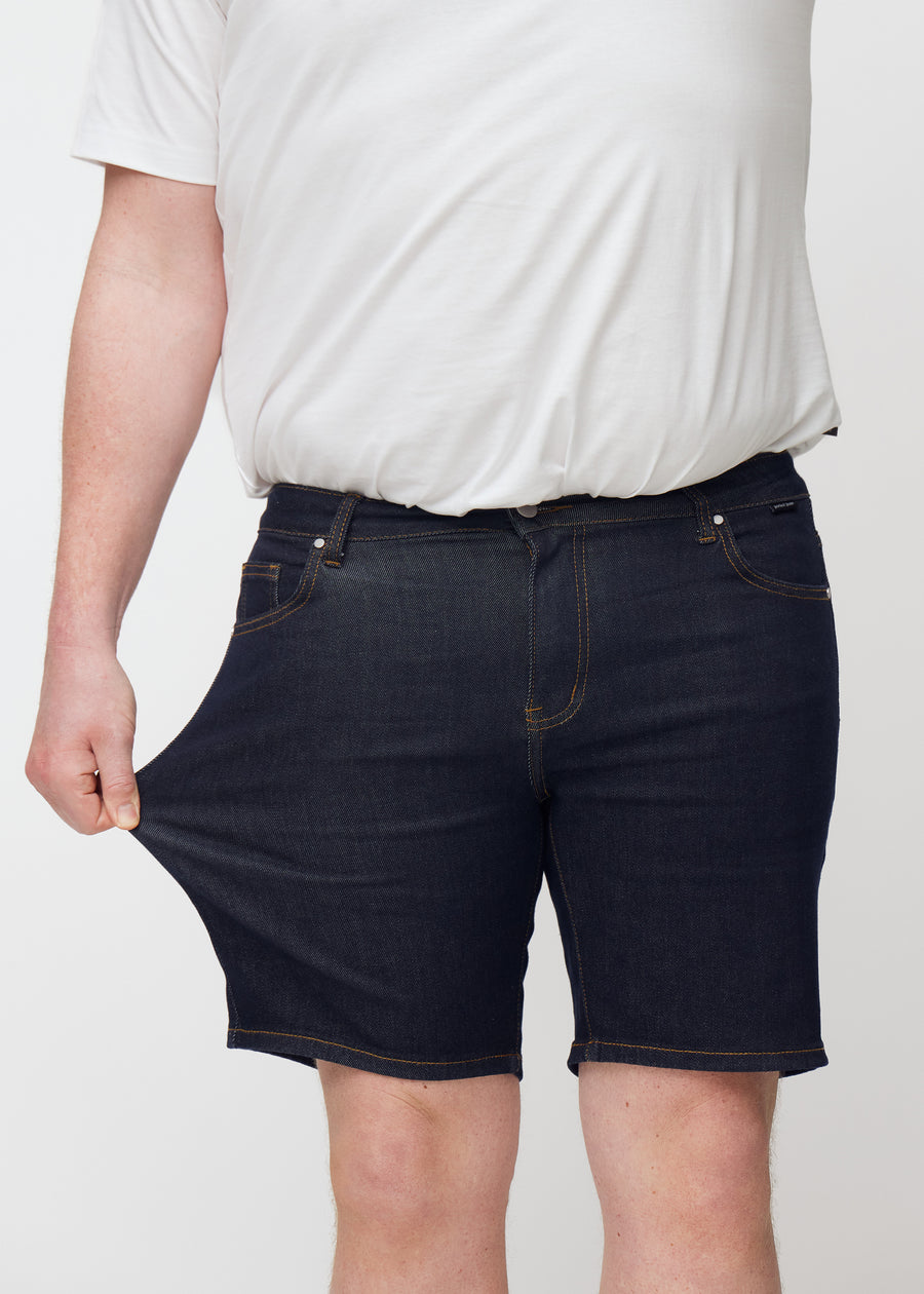 Plus-Size-Model dehnt tief dunkelblaue Denim-Shorts für Herren in der Farbe Midnights am Oberschenkel, um den Stretch zu zeigen.