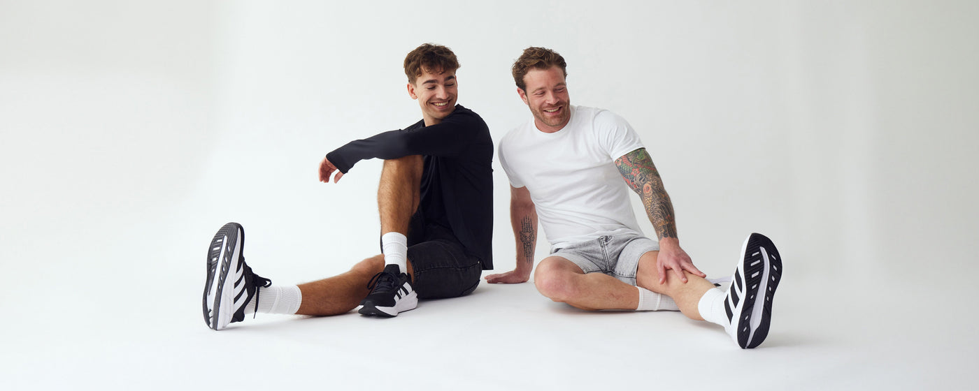 To mænd der sidder og strækker ud, iført lyse- og mørkegrå denim stretch shorts fra Perfect Jeans.